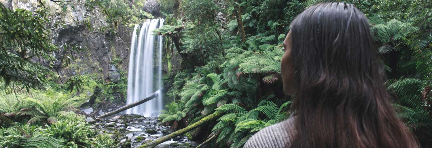 Person standing in a lush green forest looking towards a waterfall